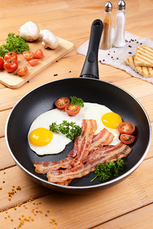 Eine Keramikpfanne mit Spiegelei und Speck auf einem Holztisch, Gewürze und Gemüse steht im Hintergrund.